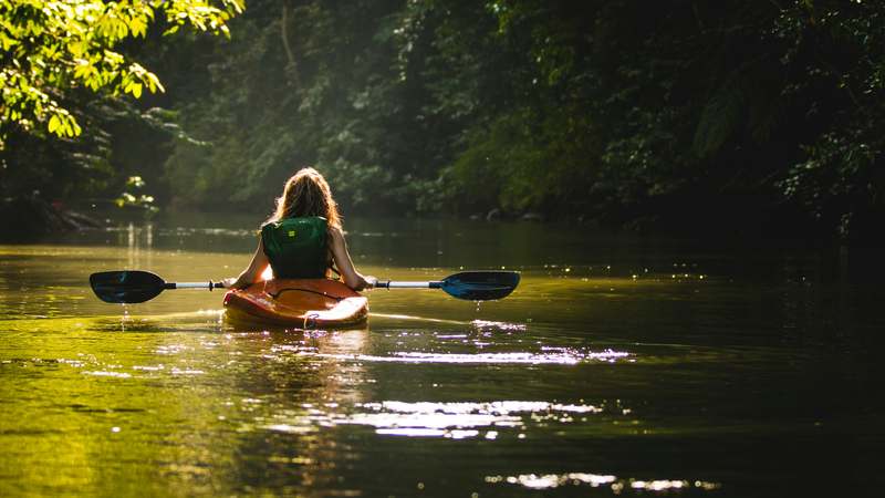A calm river with two recreational kayakers in bright yellow and blue kayaks, sunlight reflecting on water, forested shoreline in background — representing kayak safety and rental context